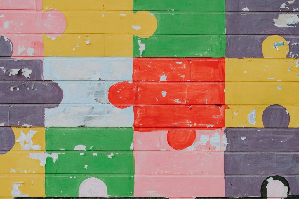 The image is a color photograph of a brick wall painted to look like a puzzle; title card for the new flash fiction, "The day they bomb the art gallery," by Belinda Rowe.