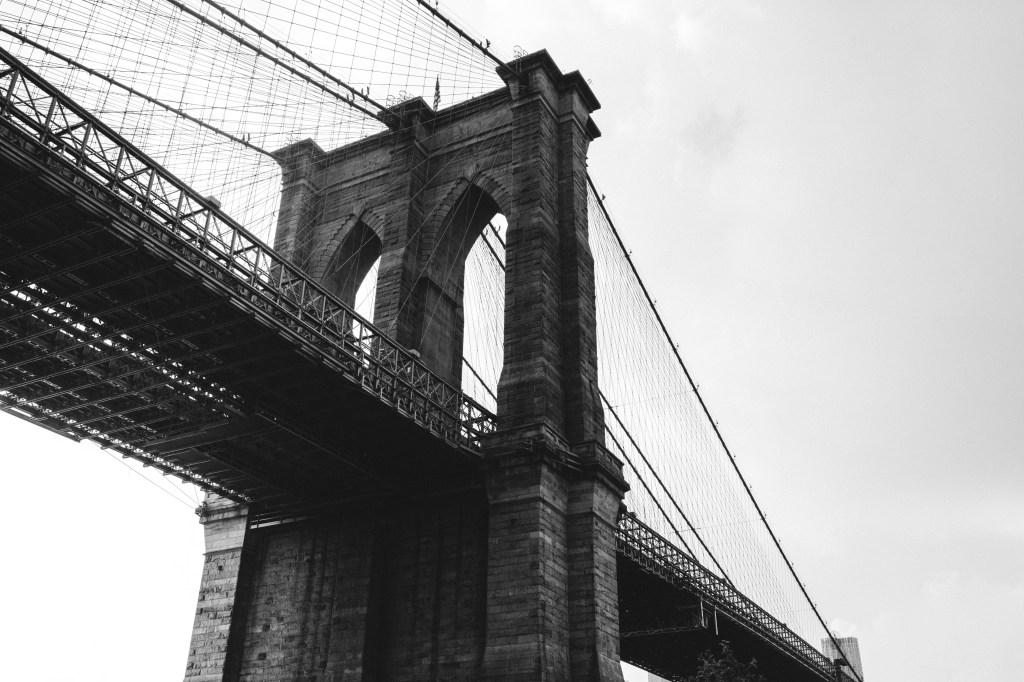 Image is a black-and-white photograph of the Brooklyn Bridge; title car for the new microfiction, "Ben Stiller’s Friend Flipped Me Off at a Restaurant in New York City,” by Rachel M. Hollis.