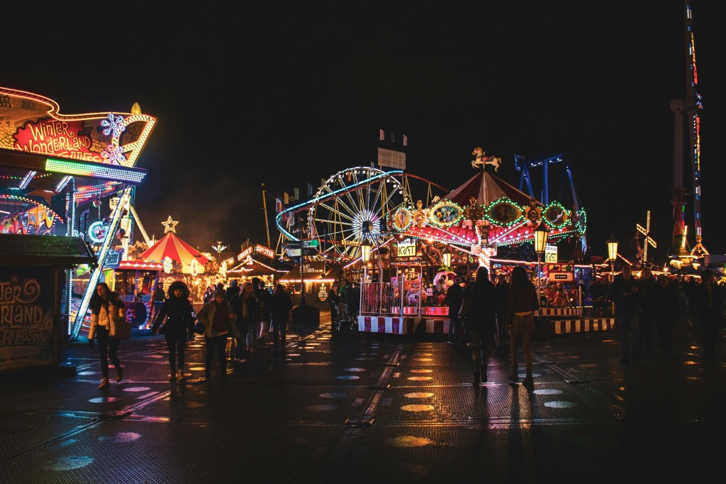 Image is a color photograph of a carnival at night; title card for the new microfiction, "Capsule," by Adele Gallogly.
