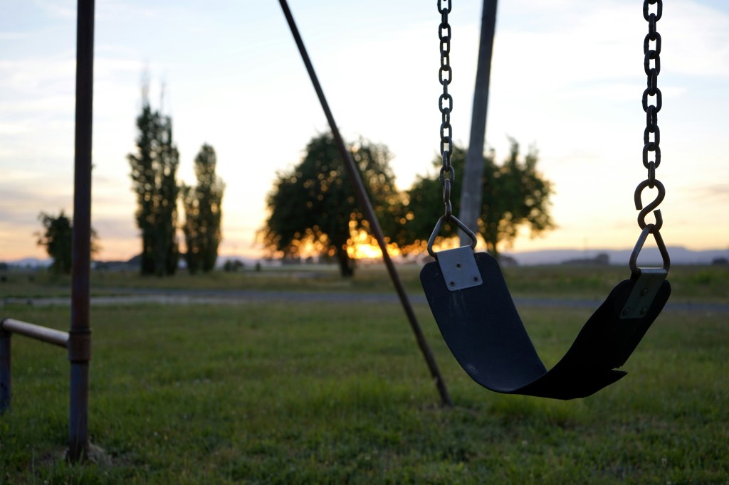 Image is a color photograph of a single swing at a small children's park; title card for the new flash fiction, "Crock Mother," by Rebecca Klassen.