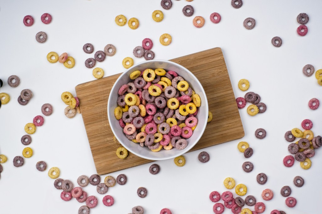 Image is a color photograph of a bowl of colorful cereal, some spilt; title card for the new flash fiction, “A Boy Wants His Mother,” by Lisa Thornton.