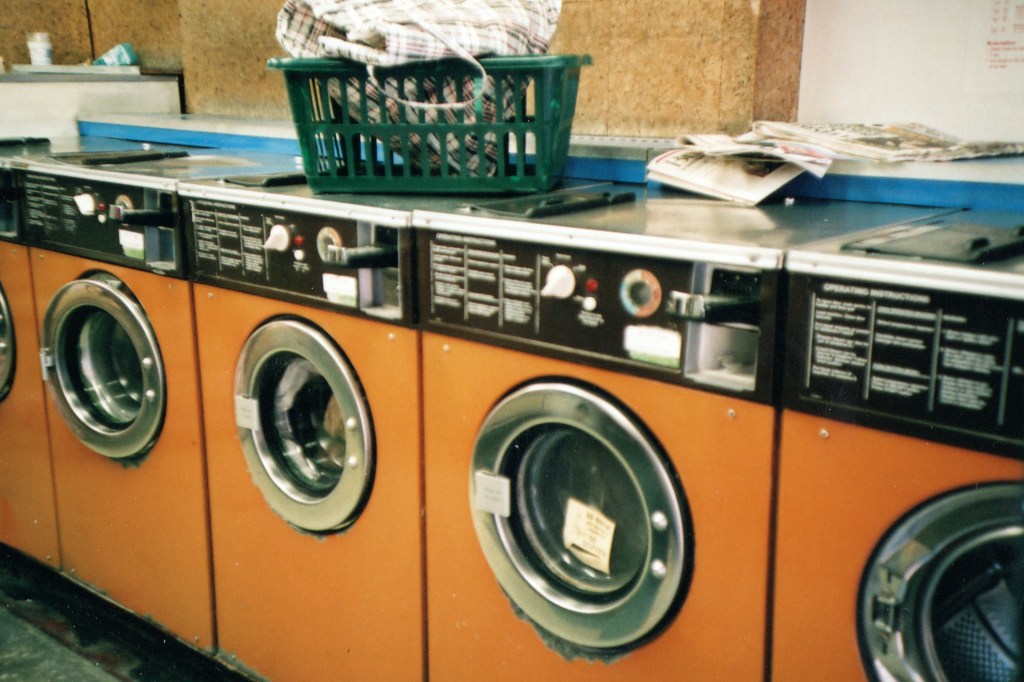 Image is a color photograph of a laundromat's orange washers and dryers; title card for the new flash fiction, “May the Circle Be,” by Maximilian Martini.